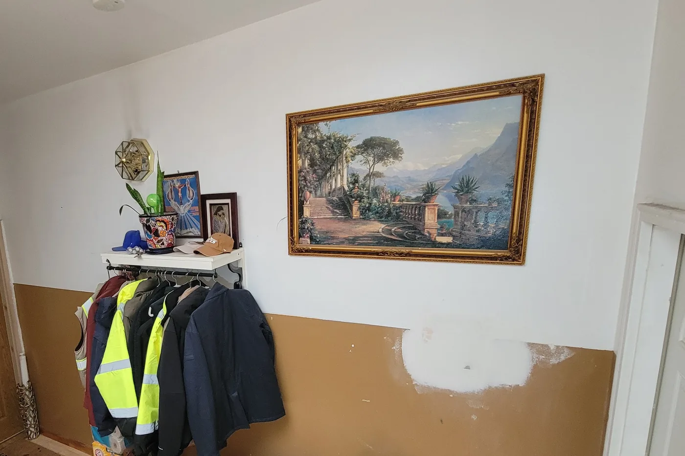 The same entry wall before painting: white above, tan below, a large bare plaster patch in the middle, coats hung in front of it.