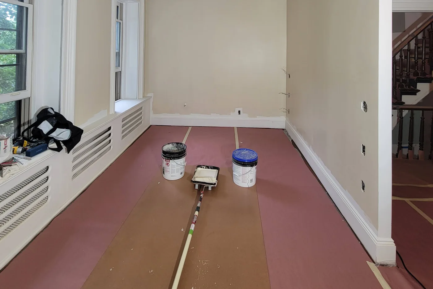 A room ready for paint: the floor covered in taped rosin paper, two buckets and a roller tray on it, fresh patches along the walls.