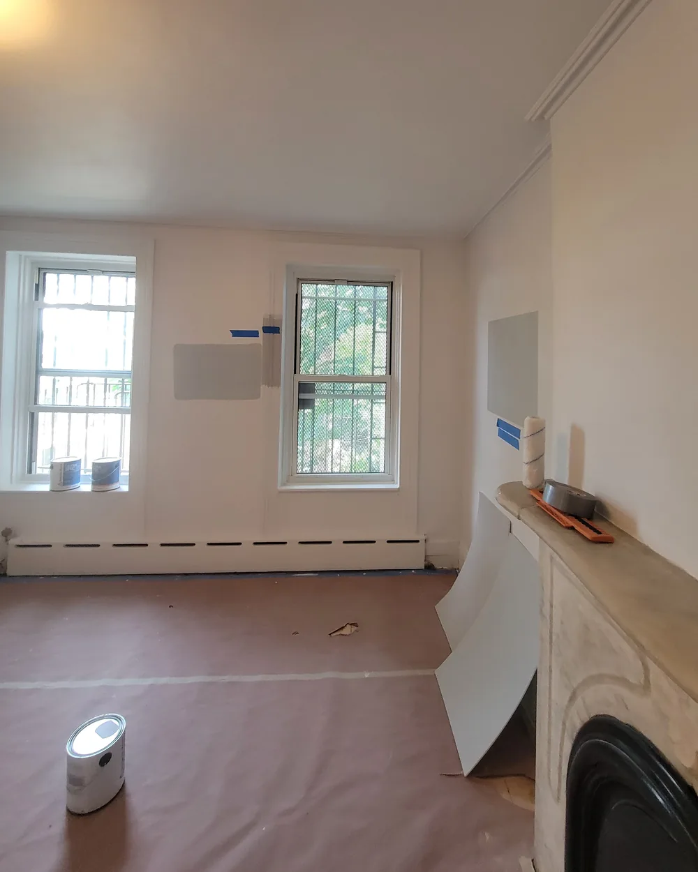 A room mid-job: paper over the floor, blue tape and gray paint test patches on white walls, a paint roller and a roll of tape on the fireplace mantel, a paint can on the floor.